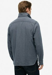 Szara bluza polarowa o teksturowanym wykończeniu, z wysokim kołnierzem, długimi rękawami i mankietami zapinanymi na guziki, charakteryzująca się luźnym krojem i szczegółami szwu w tyle.