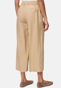 Beige, cropped wide-leg trousers with an elastic waistband and two back pockets, paired with flat, studded brown shoes. Smooth fabric texture.