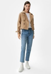 Cropped beige jacket with two front buttons and a collar, paired with light blue jeans and white sneakers. Features a classic design and fit.