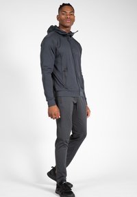 Donkergrijze sportjas voor heren met capuchon, ritszakken en lange mouwen, gecombineerd met bijpassende taps toelopende broeken en zwarte schoenen.