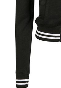 Manche de veste noire avec un bord-côte blanc rayé. Le matériau semble lisse avec une coupe ajustée et un bouton visible sur l'épaule.