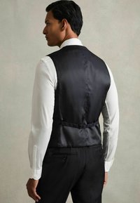 Vesta din satin negru cu o textură netedă și curea ajustabilă la spate. Purtați-o peste o cămașă albă cu mâneci lungi și pantaloni negri.