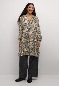 Bež tunika dugih rukava s cvjetnim uzorkom, s V-izrezom, nabranim manžetama i slobodnim krojem, u kombinaciji s tamnim hlačama širokih nogavica.