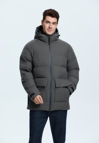Gray padded jacket with a hood, front zip closure, two large front pockets, elastic cuffs, and a smooth finish. Worn over navy jeans.