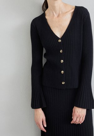 Zwarte geribbelde cardigan met een V-hals en gouden knopen. Lange mouwen met een uitlopende stijl, gecombineerd met een bijpassende geribbelde rok.