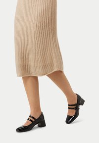 Svarta patenterade läderklackar med två smala remmar över fotryggen, rundad tå och en glänsande yta.