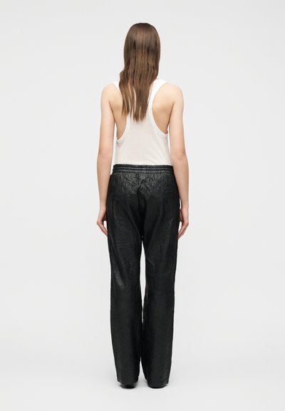 Γυναίκα με μακριά καστανά μαλλιά στέκεται γυρισμένη πλάτη, φορώντας λευκό αμάνικο τοπ και χαλαρό μαύρο υφασμάτινο παντελόνι σε απλό φόντο.