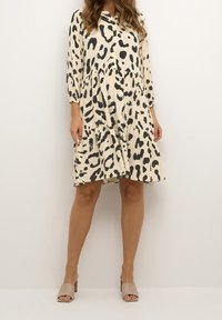 Kremowa sukienka w czarne abstrakcyjne plamy; posiada guziki z przodu, długie rękawy i falbaniaste spódnice. Połączona z beżowymi sandałami na blokowym obcasie.