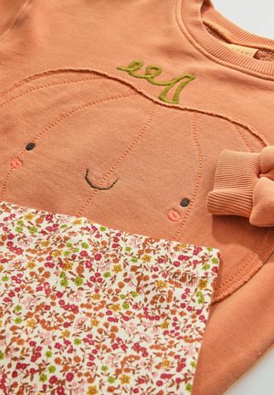 Peach sweatshirt with pumpkin face embroidery and green accents, paired with floral-patterned, ruffled, pastel fabric.