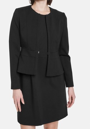 Femme portant un blazer noir ajusté à manches longues par-dessus une robe noire assortie, montrée du cou jusqu'à la mi-cuisse sur un fond uni.