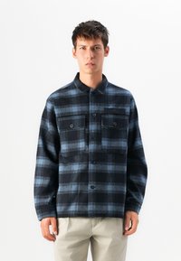 Giovane uomo con una camicia a flanella a quadri nera e blu con taschini sul petto, in piedi davanti a uno sfondo bianco uniforme.