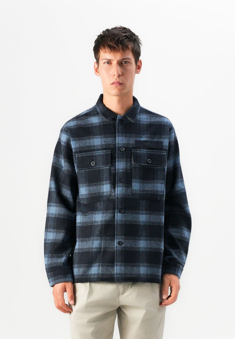 Giovane uomo con una camicia a flanella a quadri nera e blu con taschini sul petto, in piedi davanti a uno sfondo bianco uniforme.