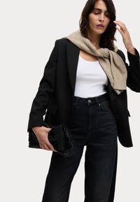 Černý blazer přes bílý top s ramínky, šedá šála přehozená, doplněno vysokými tmavými džínami. Drží strukturovanou černou kabelku.