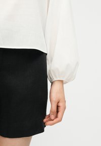 La main d'une femme pend à côté d'une jupe noire et d'un chemisier blanc à manches longues avec un poignet froncé sur un fond uni.
