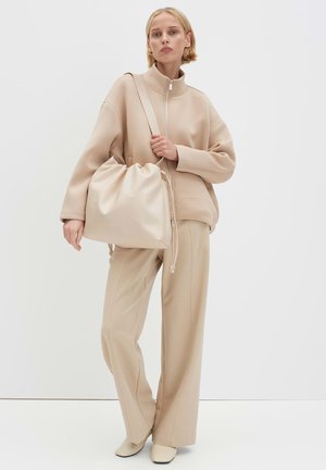 Beige drawstring bag made of smooth material, paired with a zippered beige pullover and wide-legged trousers. The outfit is monochromatic.