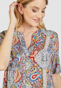 Bunte Bluse mit Blumen- und Paisleymuster, rundem Kragen und kurzen Ärmeln, die eine Mischung aus roten, blauen, gelben und weißen Details aufweist.