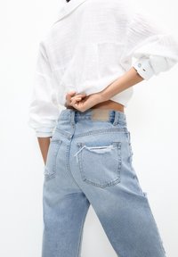 Jean en denim bleu clair avec une coupe décontractée, présentant des détails déchirés, deux poches arrière et une étiquette en cuir au niveau de la taille.