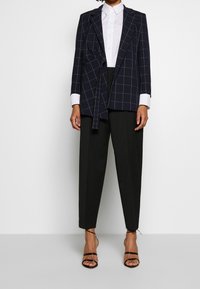 Marinblå rutmönstrad kavaj med midjeband, vit skjorta med knappar och svarta skräddarsydda byxor; tillsammans med svarta sandaler med remmar.