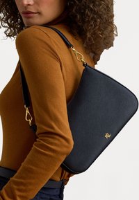 Μαύρη τσάντα ώμου με υφή, με χρυσό λογότυπο και χρυσές λεπτομέρειες, διαθέτει φερμουάρ και καμπυλωτό σχήμα. Φοριέται πάνω από ένα καφέ μπλουζάκι.