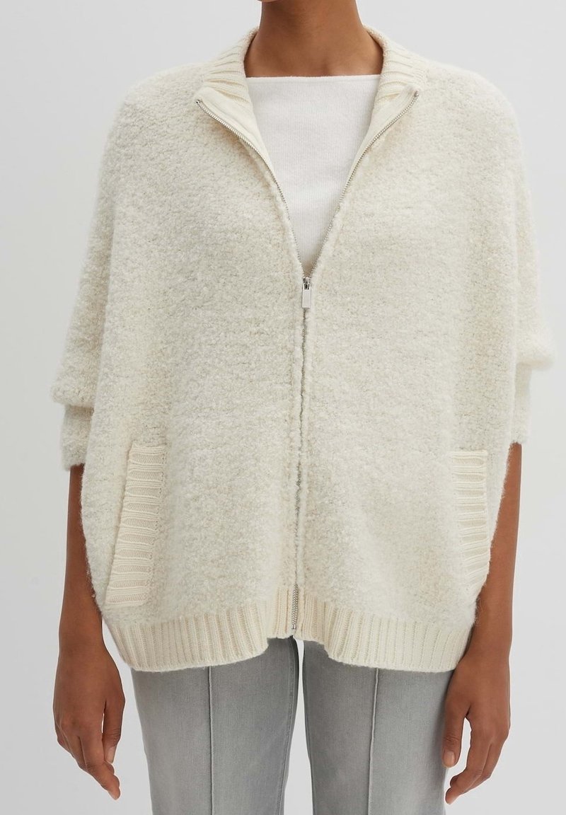 Kermanvärinen vetoketjullinen neuletakki, valmistettu pehmeästä, teksturoidusta kankaasta. Siinä on resorireunaiset hihansuut, kaksi sivutaskua ja rento istuvuus.