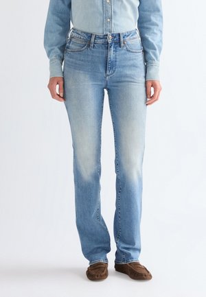 Kobieta ubrana w jasnoniebieskie dżinsy i pasującą dżinsową koszulę, stojąca z rękami swobodnie opuszczonymi wzdłuż ciała, w brązowych mokasynach.