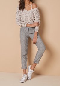 Lichtroze gebreide trui met open weving, gecombineerd met grijze taps toelopende broeken en witte sneaker. De trui heeft een off-shoulder ontwerp en lange mouwen.