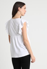 Top de algodão branco com mangas em babados, cintura franzida e textura suave. Usado com calças pretas, mostrando um estilo casual.