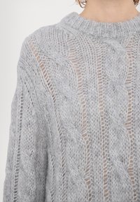 Gros plan sur une personne portant un pull en maille torsadée gris clair, ample, avec un col rond côtelé.