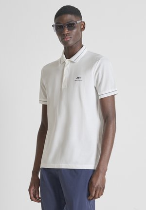 Hombre con gafas de sol negras, camisa polo blanca con logo y pantalones azul marino, de pie contra un fondo claro y liso.