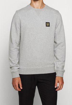 Mann trägt hellgrauen Langarm-Sweatshirt mit kleinem schwarzem und goldenem Logo-Patch auf der Brust, kombiniert mit schwarzen Hosen.