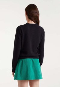 Schwarzer Pullover mit langen Ärmeln und Rundhalsausschnitt, kombiniert mit einem grünen Cordrock. Glattes Material und taillierte Passform. Rückansicht.
