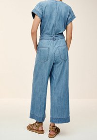 Combinaison en denim bleu clair avec manches courtes, taille ceinturée, jambes larges et deux poches arrière, associée à des sandales marron à lanières.