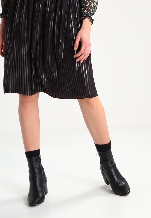 Jupe noire plissée avec une finition brillante, assortie à des bottines noires. La tenue met en avant des lignes verticales et un tissu texturé.
