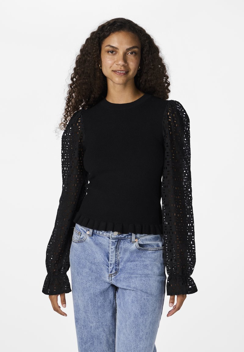 Maglione nero a maglia con orlo arricciato e maniche a sbuffo in pizzo nero trasparente, caratterizzato da un motivo a ritaglio geometrico. Indossato con jeans azzurri chiari.