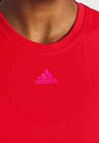 Primer plano del cuello y la parte superior del pecho de una persona que lleva una camiseta deportiva roja de Adidas con un logo rosa.
