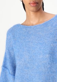 Απαλή μπλε πλεκτή μπλούζα με φαρδύ λαιμό και χαλαρή εφαρμογή. Η υφή είναι μαλακή, με ελαφριά, ελαφρώς φουντωτή φινέτσα.