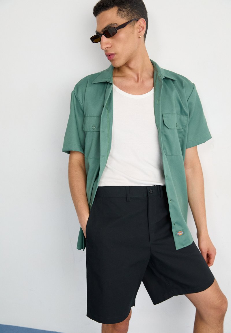 Kortärmad grön skjorta med knappar över en vit linne, kombinerat med svarta shorts. Funktioner inkluderar bröstfickor och en logotyp från märket på shortsen.