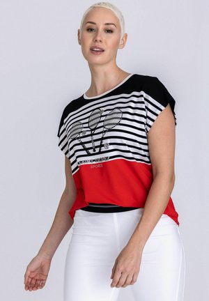 Femme portant un haut à rayures noires, blanches et rouges avec des graphismes de raquettes de tennis et un pantalon blanc, debout devant un fond uni.