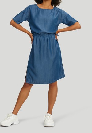 Žena v midi modrých džínových šatech s krátkými rukávy a bočními rozparky, obutá v bílých robustních teniskách, s rukama v bok proti jednoduchému pozadí.