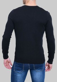 Pull en tricot noir à manches longues, col rond et ourlet côtelé. Porté par-dessus un jean bleu, mettant en évidence une texture lisse et un design ajusté.