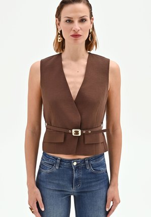 Donna con gilet marrone senza maniche con cintura e jeans blu, con orecchini dorati e capelli corti, in piedi davanti a uno sfondo chiaro e uniforme.