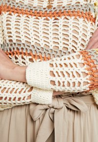 Pull en crochet ivoire, gris et orange avec un tissage ouvert. Manches longues avec poignets côtelés. Jupe beige à nouer à la taille en dessous.