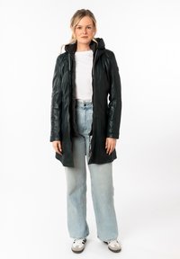 Chaqueta de cuero negra con capucha, que presenta un cierre delantero, mangas ajustadas y una textura sutil. Lleva puesta sobre una camisa blanca y jeans de mezclilla azul.
