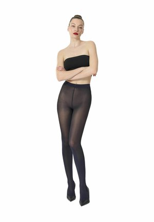 Frau steht mit verschränkten Armen, trägt ein schwarzes Bandeau-Top, transparente schwarze Strumpfhosen und schwarze High Heels vor weißem Hintergrund.
