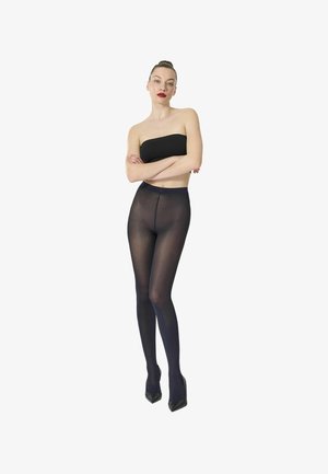 Frau steht mit verschränkten Armen, trägt ein schwarzes Bandeau-Top, transparente schwarze Strumpfhosen und schwarze High Heels vor weißem Hintergrund.