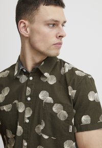 Camisa de manga corta en color verde oliva, fabricada en tejido ligero con estampado de conchas blancas, que cuenta con cierre de botones en la parte delantera y un bolsillo en el pecho.