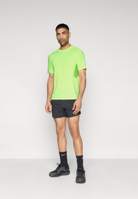 Αθλητική μπλούζα σε νέον πράσινο χρώμα με κοντά μανίκια και λογότυπο, συνδυασμένη με μαύρο σορτς, μαύρα αθλητικά παπούτσια και μαύρες κάλτσες. Λείο ύφασμα και εφαρμοστή γραμμή.