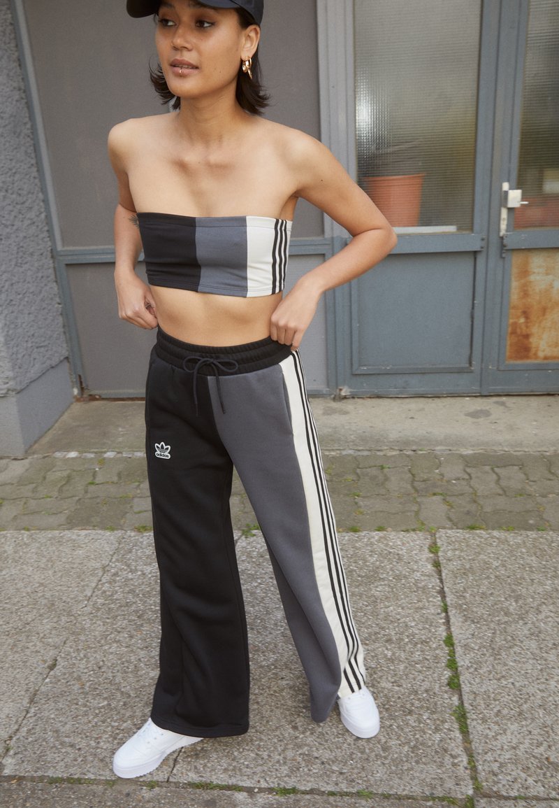 Čierny a šedý farebne delený crop top a zodpovedajúce široké nohavice s bielymi pruhmi a logom. Model má na sebe biele tenisky a čiernu šiltovku.