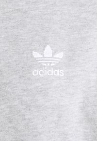 Vitt Adidas trefoil-logotyp broderad på ljusgrått tyg, som visar varumärkets emblem centrerat på textilytan.