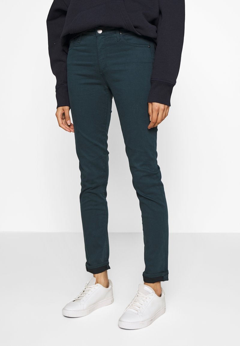 Dark teal, slim-fit pants with a smooth texture, front pockets, and a single button closure, paired with white sneakers.